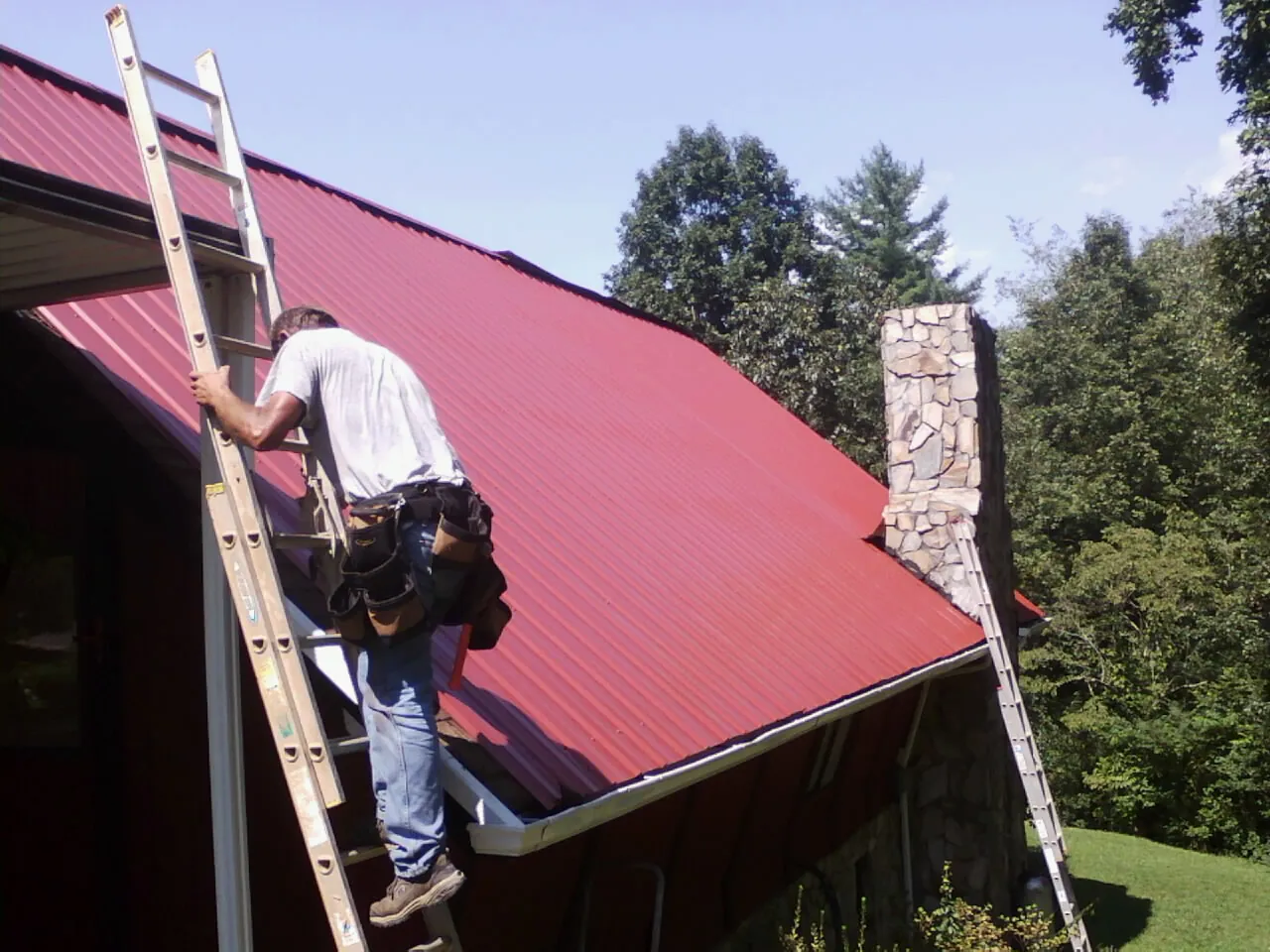 Roofer on a ladder beside a red metal roof and stone chimney — Soffit & Fascia Repair by Roofing Brothers in Sea Isle City